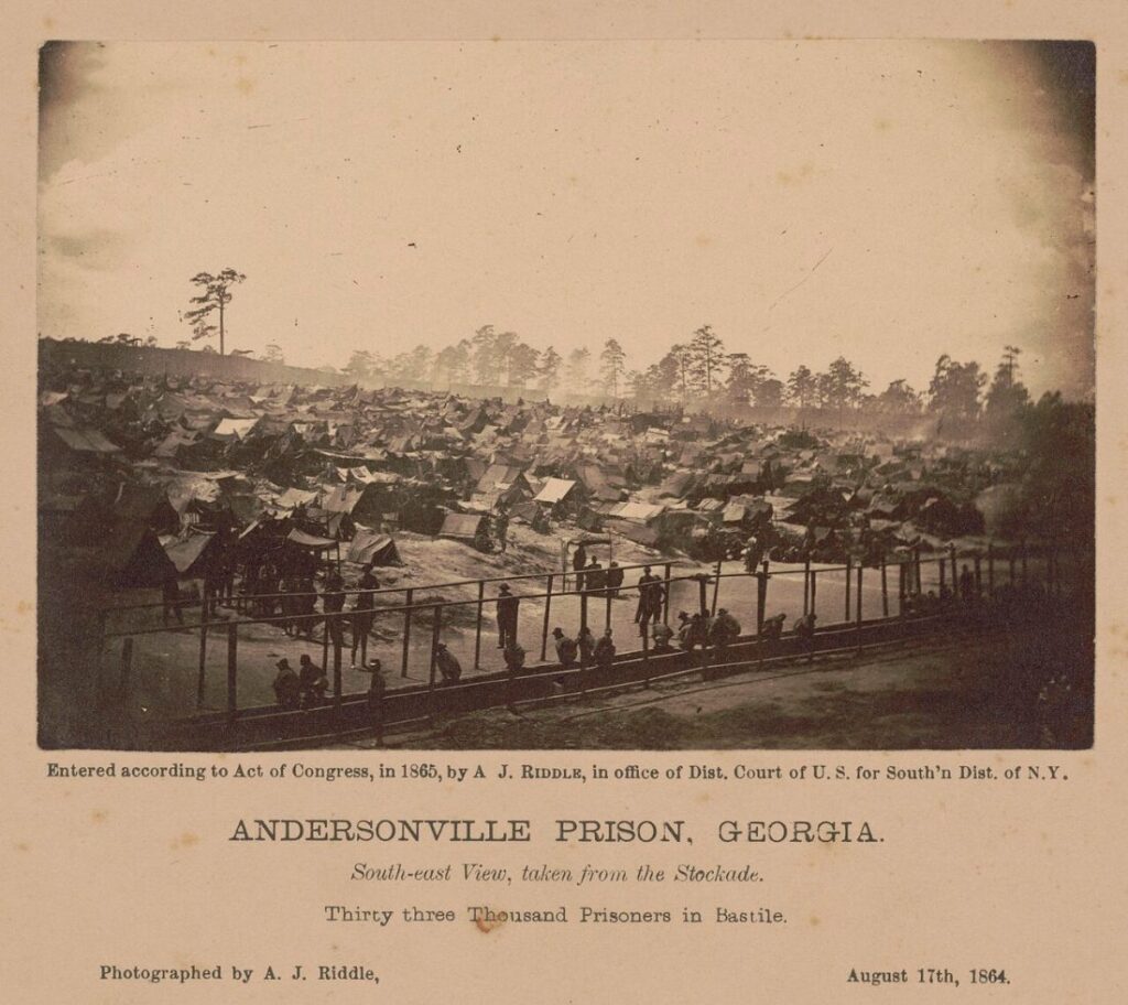 An 1864 albumen photograph looking south-east across Andersonville prison, showing a hillside covered edge to edge with improvised tents and shelters behind the stockade rail