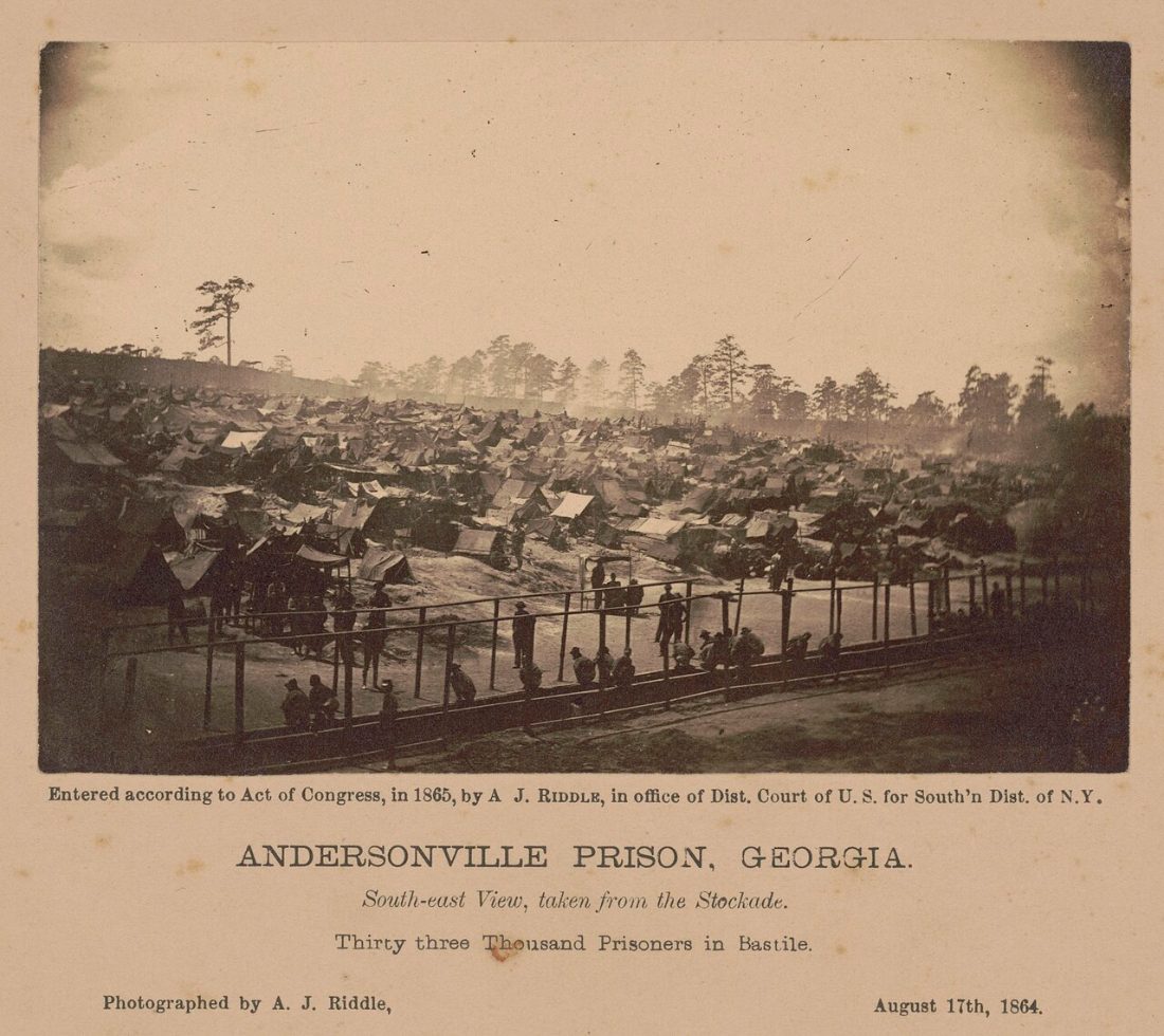An 1864 albumen photograph looking south-east across Andersonville prison, showing a hillside covered edge to edge with improvised tents and shelters behind the stockade rail