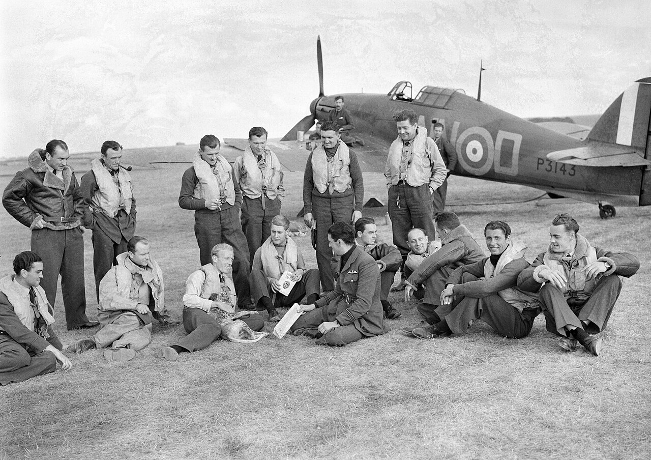 A line of pilots in flying kit sitting and standing in front of a Hurricane fighter