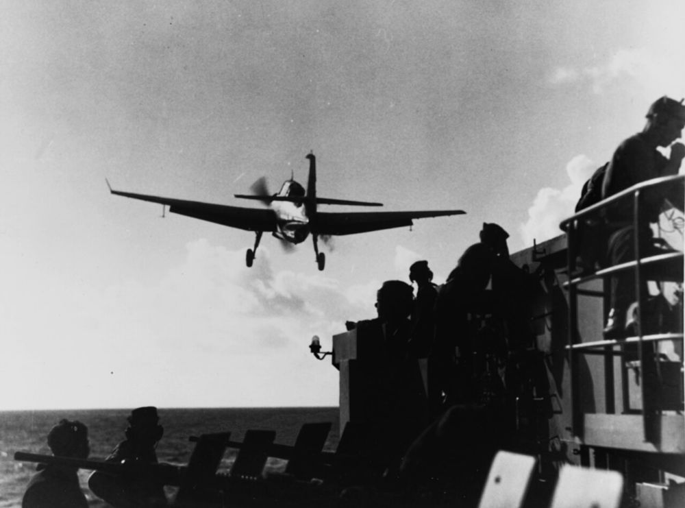 A TBM Avenger with landing gear down silhouetted above the deck of an aircraft carrier, with crewmen in the foreground