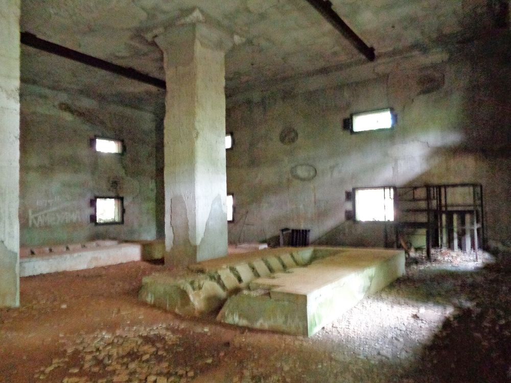The bare concrete interior of a ruined wartime building on Chichijima, with a central pillar, small windows and equipment plinths