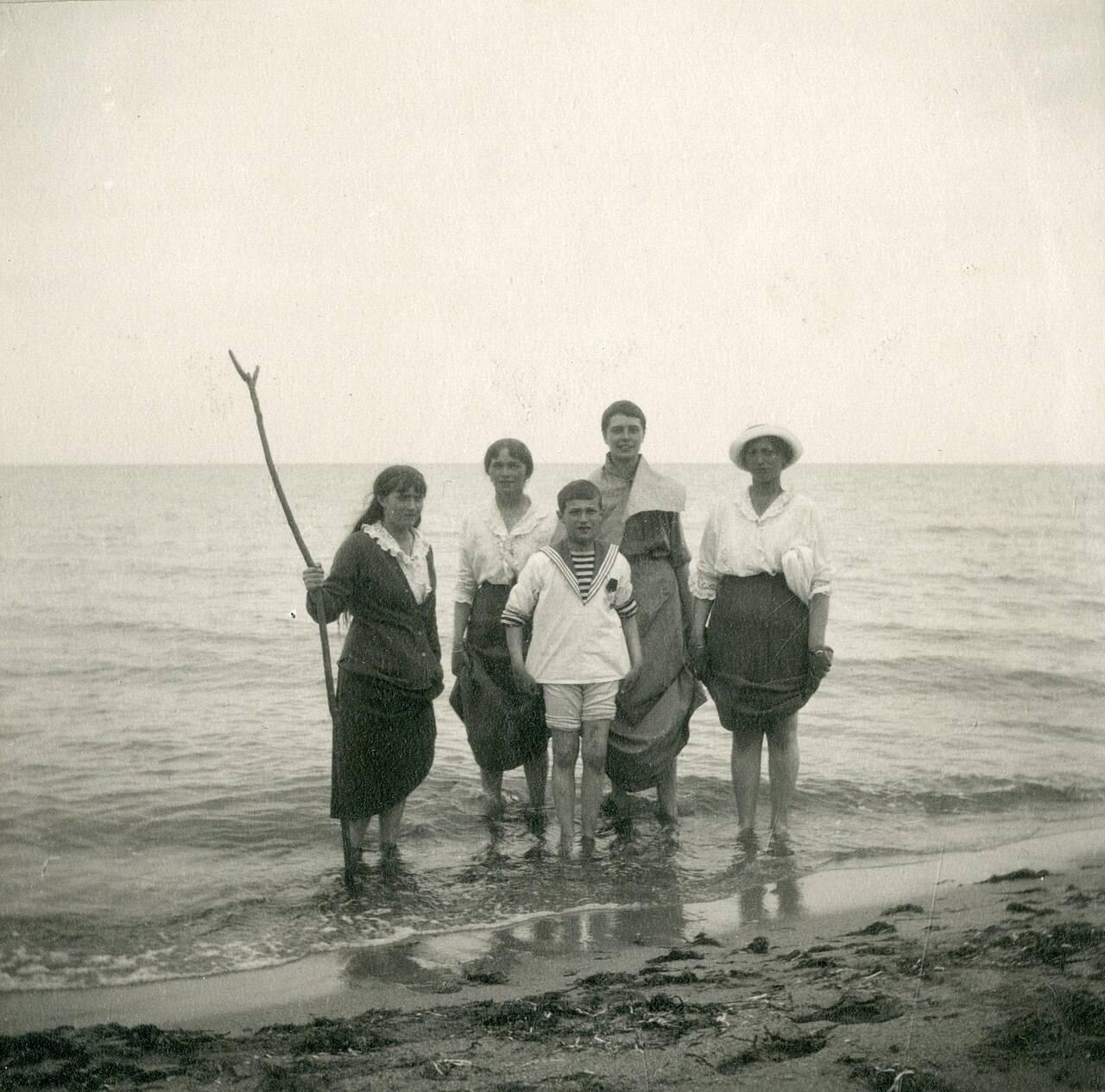 A group standing in the shallows at a beach, holding up their skirts