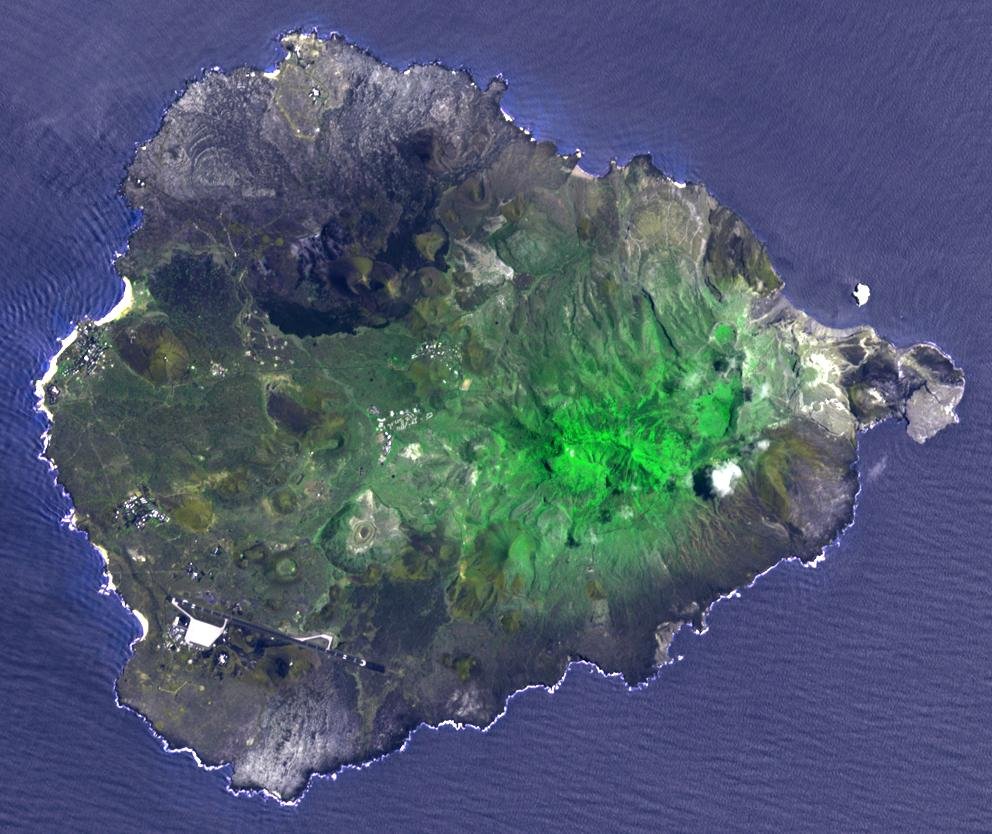 A satellite image of Ascension Island, an isolated volcanic island in dark grey and brown surrounded by deep blue ocean, with a patch of bright green at its centre