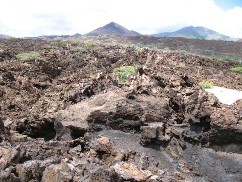 A broken field of black and rust-coloured lava rock on Ascension Island, with volcanic cones rising in the distance