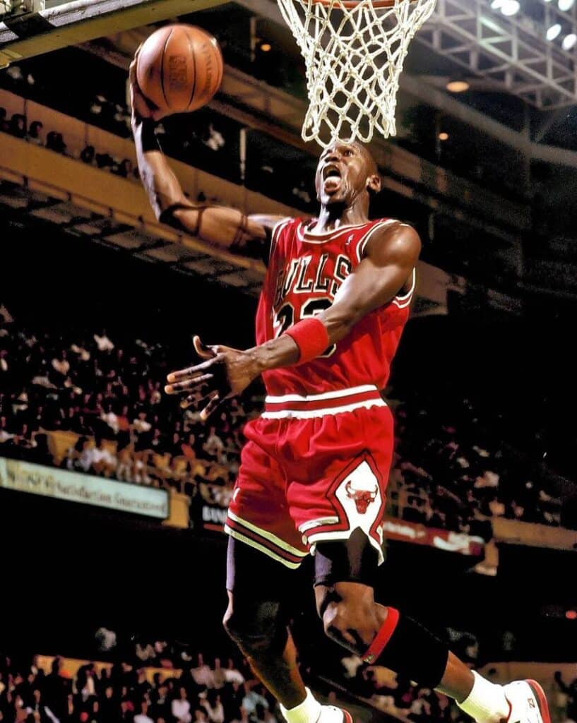 Basketball player in a red Bulls uniform slam dunks toward the basket in a crowded arena.