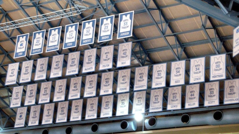 Rows of North Carolina Tar Heels jersey banners hanging from an arena ceiling, with Jordan's number 23 among the retired numbers in the top row