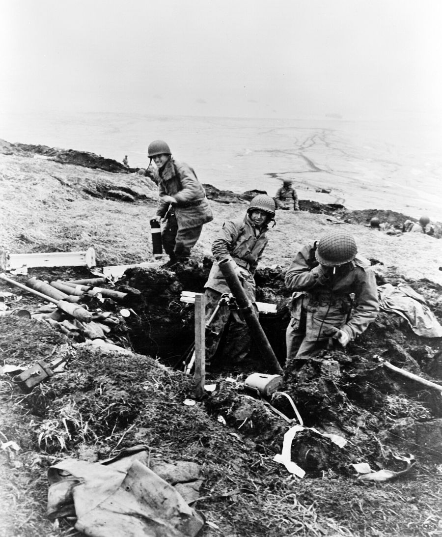 Three American soldiers in a muddy pit on a bare ridge load a trench mortar, with shells stacked beside them, Attu 1943