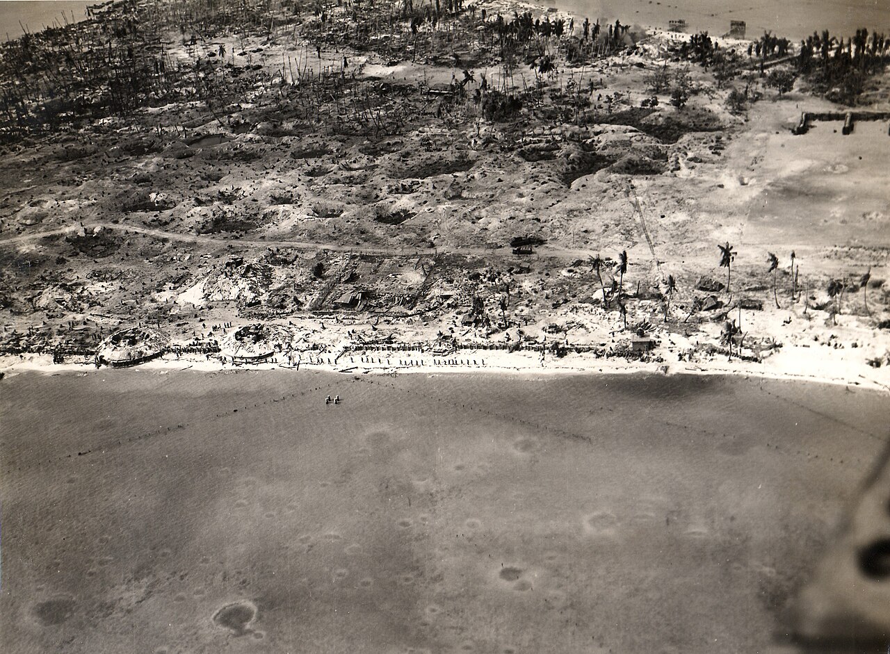 Low aerial view of a narrow, flat island scarred by shellfire and littered with wreckage