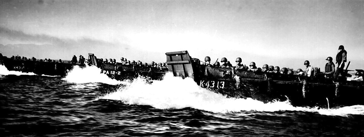A long line of landing barges crossing open water toward a shore