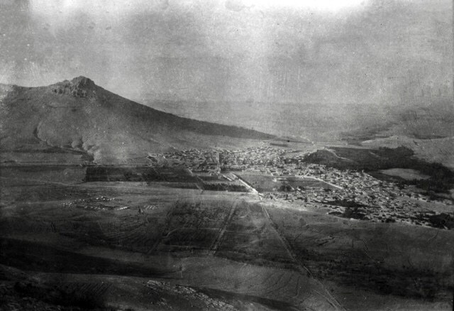 A hazy black and white view of the Persian town of Bijar spread across a valley floor beneath a bare hill, photographed from high ground
