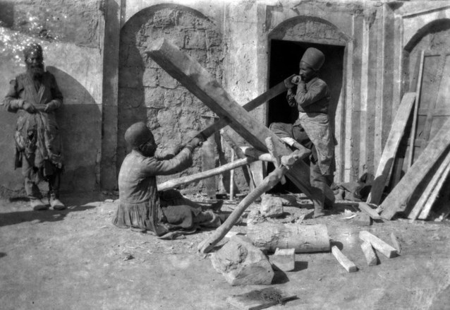 Two men sawing a poplar trunk into planks in a courtyard while an older bearded man in ragged clothing looks on