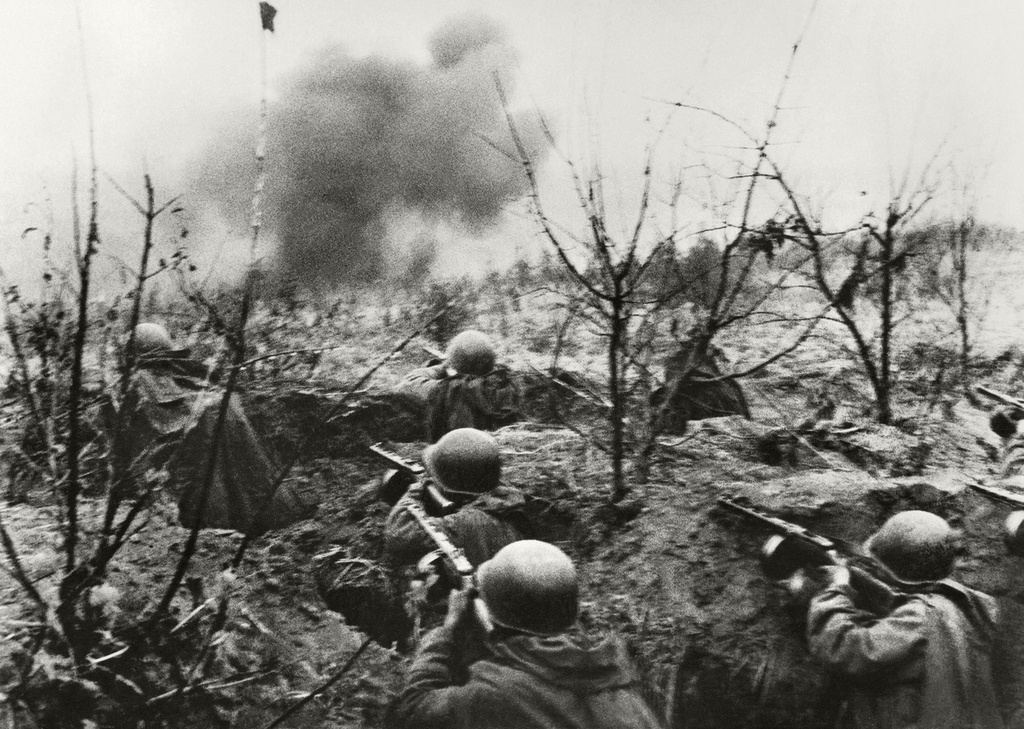 Soldiers with submachine guns in a shallow trench cut through rubble, smoke behind them