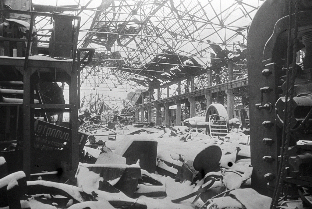 The gutted interior of a factory hall, roof trusses collapsed, snow over the machinery