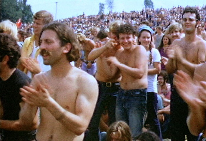 A dense crowd of young people applauding on a grassy hillside at the Woodstock festival in 1969, many shirtless in the sun