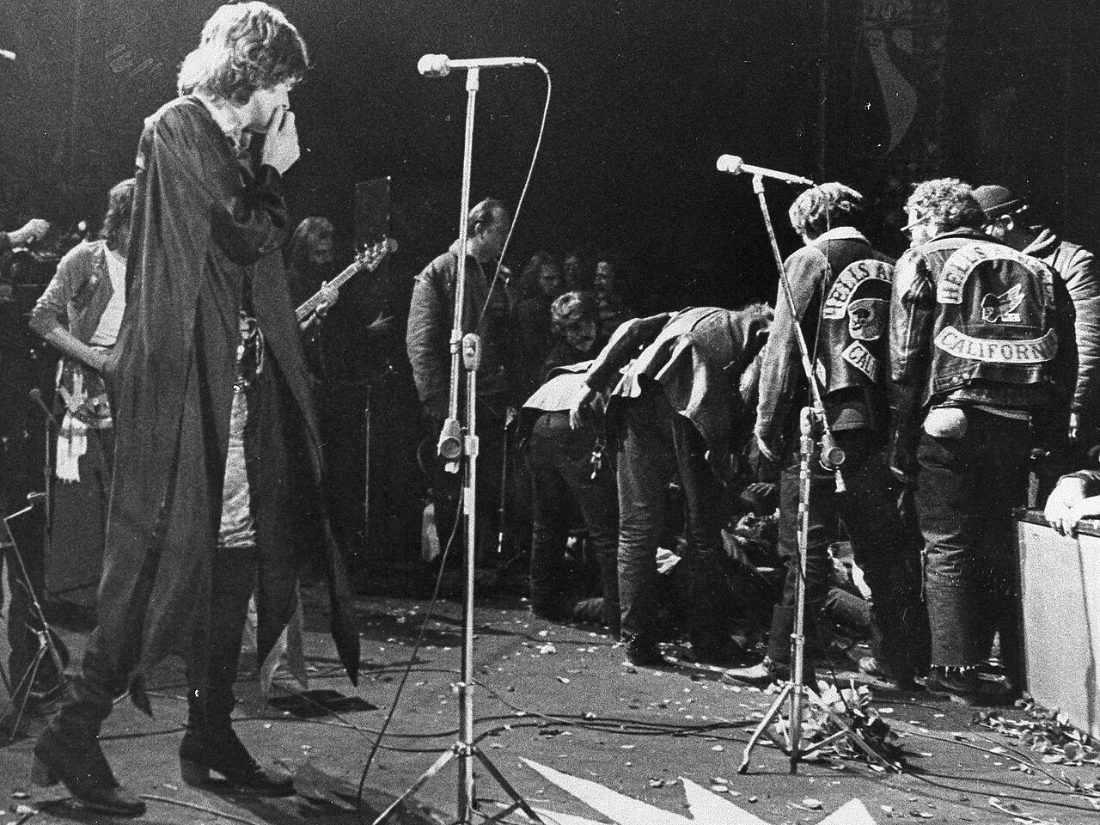 A black and white photograph of Mick Jagger standing at a microphone on the Altamont stage in 1969, watching Hells Angels in club jackets crowd the stage edge